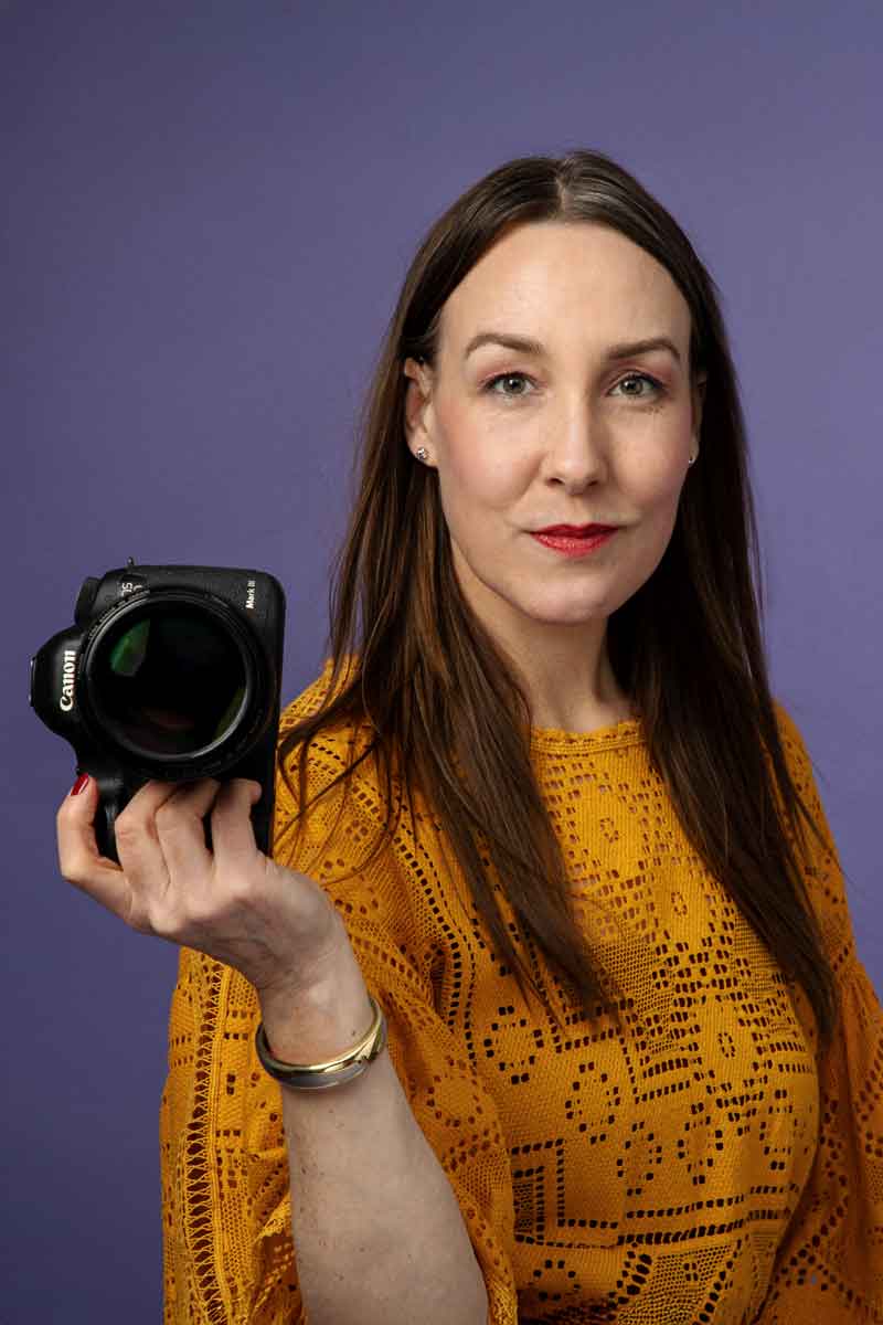 DagmarFotografinHamburg Fotografin mit orange-gelben Oberteil mit Kamera in der Hand vor lila Hintergrund